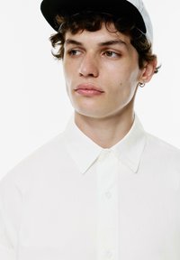 Camicia bianca con colletto a punta e maniche corte. Il modello indossa un berretto nero con la parte interna bianca e ha un orecchino.