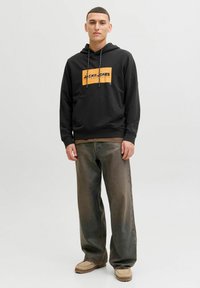 Jeune homme debout portant un sweat à capuche noir avec un logo orange, un jean gris délavé large et des mocassins marron clair.
