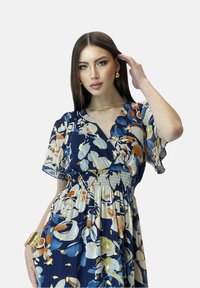 Abito floreale con sfondo blu navy profondo, caratterizzato da fiori crema, arancione e blu. Maniche corte fluenti e dettaglio in vita arricciato.