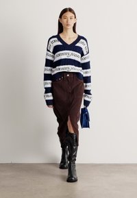 Maglione a righe navy e bianche con testo "tommy jeans", abbinato a una gonna in corduroy marrone con spacco frontale. Stivali neri al ginocchio e una borsa blu.