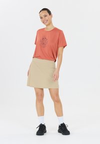 Baumwoll-T-Shirt in Rostfarbe mit grafischem Druck, kombiniert mit einem hellbeigen knielangen Rock. Schwarze Sportschuhe mit weißen Socken. Minimalistisches Design.