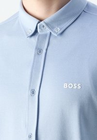Jasnoniebieska bawełniana koszula na guziki z fakturowanym wzorem, klasycznym kołnierzem i białym logo 'BOSS' na piersi, z przezroczystymi guzikami.