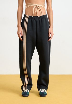Persoon draagt zwarte broek met brede pijpen en beige verticale strepen en marineblauwe sneakers op een gele vloer tegen een witte achtergrond.