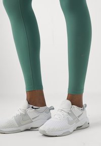 Gröna sportleggings med en slät yta och en grå Nike-logotyp på sidan, kombinerat med vita och grå Nike-sneakers med nätakenter.