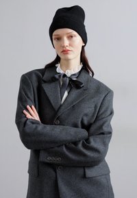 Šedý oversized blazer vyrobený z plstenej vlny, s veľkými klopami a tromi gombíkmi. Čierny čiapka a volánový biely golier dopĺňajú vzhľad.