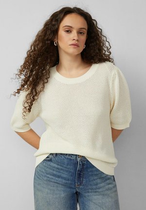 Frau mit langen lockigen Haaren, die silberne Creolen, einen weißen, strukturierten Kurzarmpullover und blaue Jeans trägt, steht vor einem grauen Hintergrund.