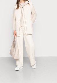 Gilet matelassé beige avec fermeture éclair à l'avant, associé à un pull beige texturé et un pantalon à jambe large ; tenant un sac beige clair, portant des baskets blanches.