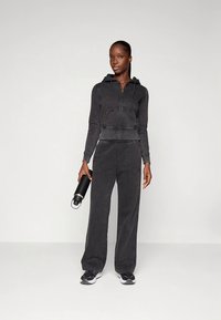 Casaco preto curto com fecho de correr, detalhes de cordão e calças largas combinando. Modelo segura uma garrafa de água em aço inoxidável.