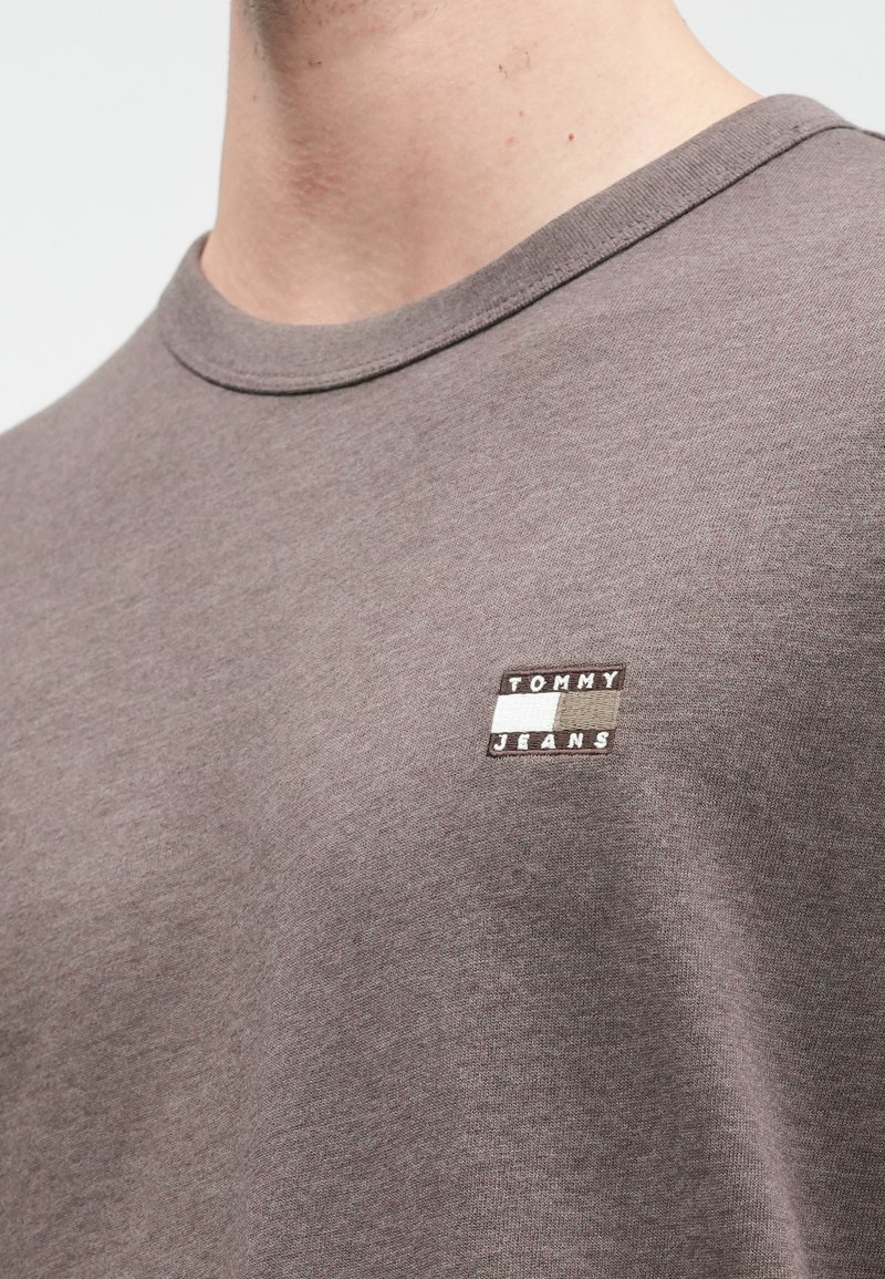 Nærbillede af en taupe crewneck t-shirt med et lille broderet Tommy Jeans-logo på brystet, båret af en person.