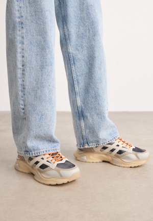 Deportivas que combinan materiales en tonos gris, negro y crema. Se caracterizan por paneles texturizados y cordones naranjas, con una suela gruesa.
