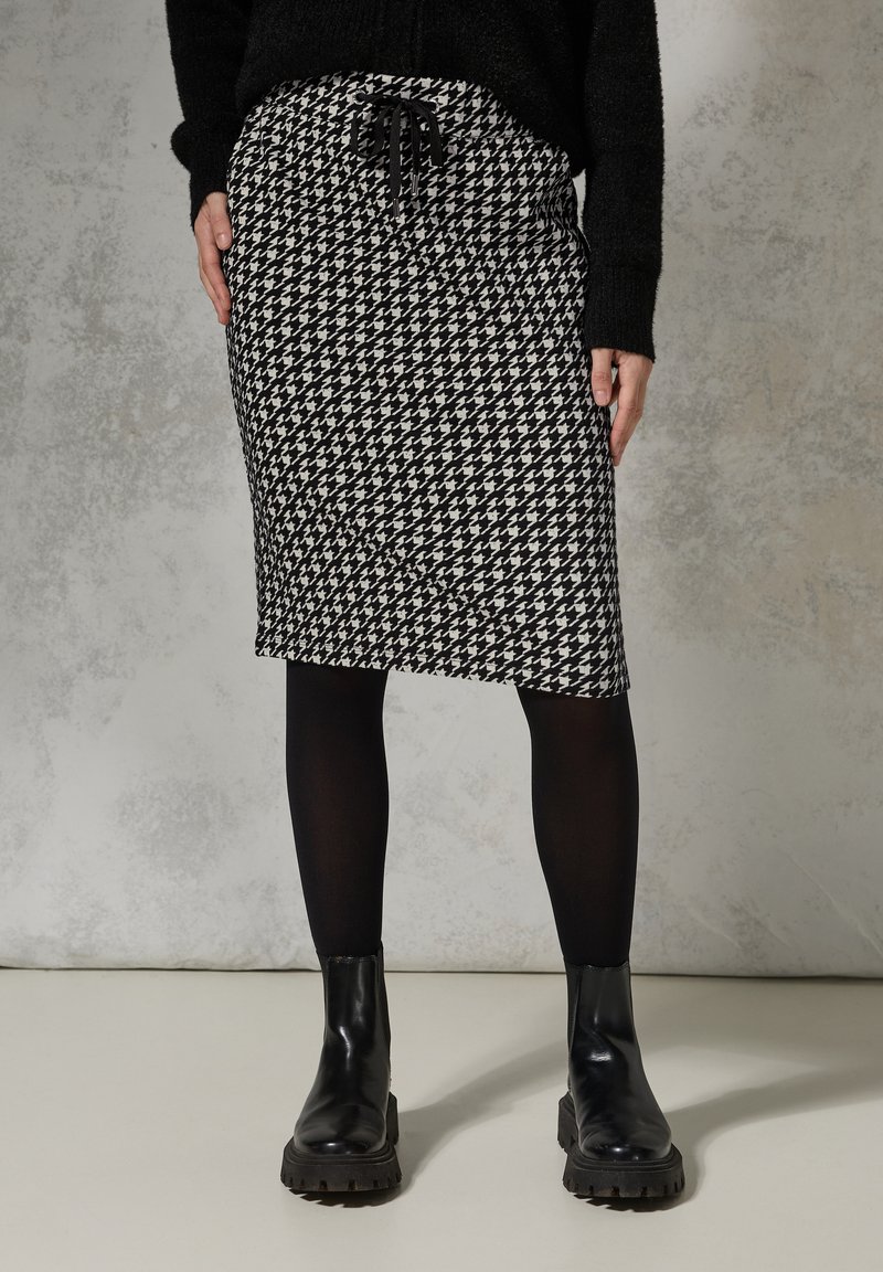 Knielange Rock im schwarz-weißen Hahnentrittmuster mit einem Kordelzugbund, kombiniert mit schwarzen blickdichten Strumpfhosen und klobigen schwarzen Stiefeln.