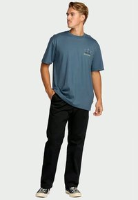 Jonge man in een blauw graphic t-shirt, zwarte broek en zwarte sneakers, staand met één hand in zijn zak, kijkend naar links.