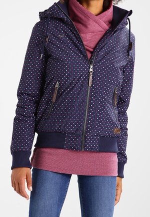 Kvinde iført navyblå lynlåsjakke med lyserøde prikker, lagdelt over en lyserød top og kombineret med blå jeans.