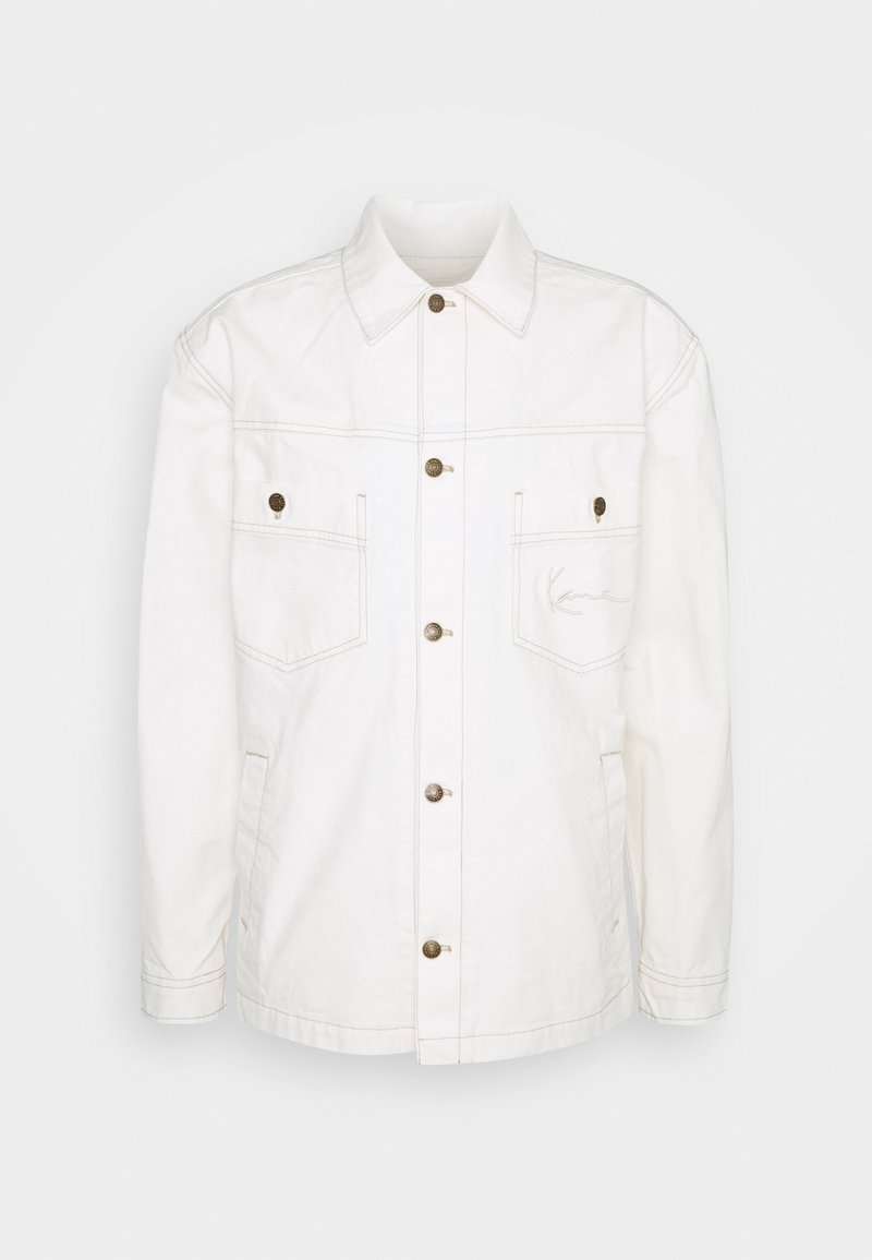 Veste blanche à manches longues boutonnée avec col, deux poches poitrine à boutons et poches latérales, présentant des détails de couture visibles.