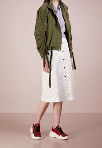 Olivgrön bomberjacka med samlade sidor, vit knälång kjol med marinblåa prickar, och röda rutiga sneakers med vita sulor.