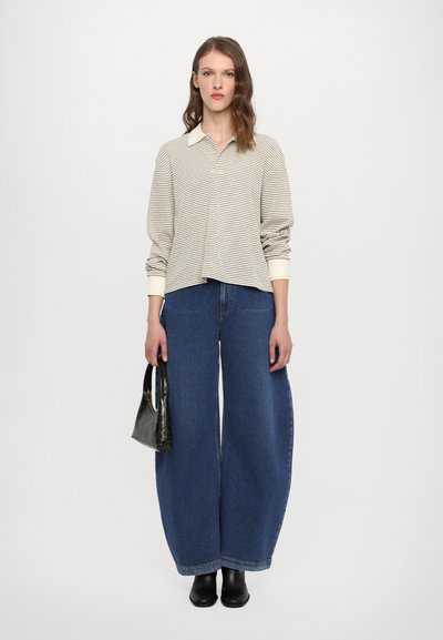 Polo a maniche lunghe a righe in crema e beige, abbinato a jeans in denim blu a gamba larga. Il modello tiene in mano una borsa nera in stampa cocco.