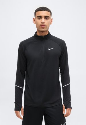 Mand iført sort Nike sportstrøje med lange ærmer, kvart lynlås og reflekterende striber på ærmerne, stående mod en ensfarvet baggrund.