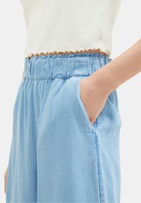 Blauwe chambraybroek met een elastische tailleband en zijdelingse zakken; de stof heeft een zachte, gladde textuur en een iets relaxte pasvorm.