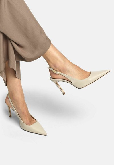Füße in beige spitz zulaufenden Slingback-Pumps, kombiniert mit lockeren braunen Hosen vor schlichtem Hintergrund.