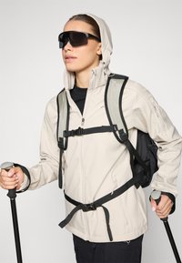 Veste softshell beige clair avec capuche, sangles noires et un sac à dos. Des lunettes de soleil foncées et des bâtons de trekking complètent l'équipement de plein air.