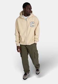 Felpa beige con zip, caratterizzata da un grande logo 'C', abbinata a pantaloni cargo verdi e sneaker neutre con suole in rete e gomma.