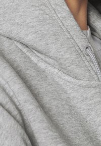 Sweat à capuche gris clair avec une texture douce, présentant un col en V, un cordon de serrage réglable et des coutures contrastantes le long des coutures.