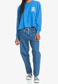 Niebieska krótka bluza z długimi rękawami i okrągłym dekoltem, zestawiona z jeansami o wysokim stanie i luźnym kroju oraz czarno-białymi trampkami.