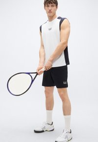 Pánske atletické tielko bez rukávov v bielej farbe s námorníckymi doplnkami, v kombinácii s námorníckymi šortkami. Drží čiernu tenisovú raketu, nosí biele tenisky a ponožky.