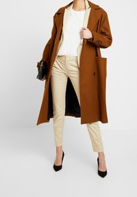 Manteau en laine marron avec revers beiges, chemise crème et pantalon beige coupe slim ; associé à des talons pointus noirs et une pochette noire.