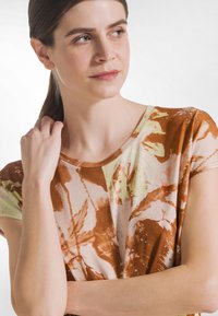 Krótki rękaw topu w mieszance brązowych i pastelowych kolorów z kwiatowym wzorem. Posiada okrągły dekolt i miękką, gładką teksturę materiału.