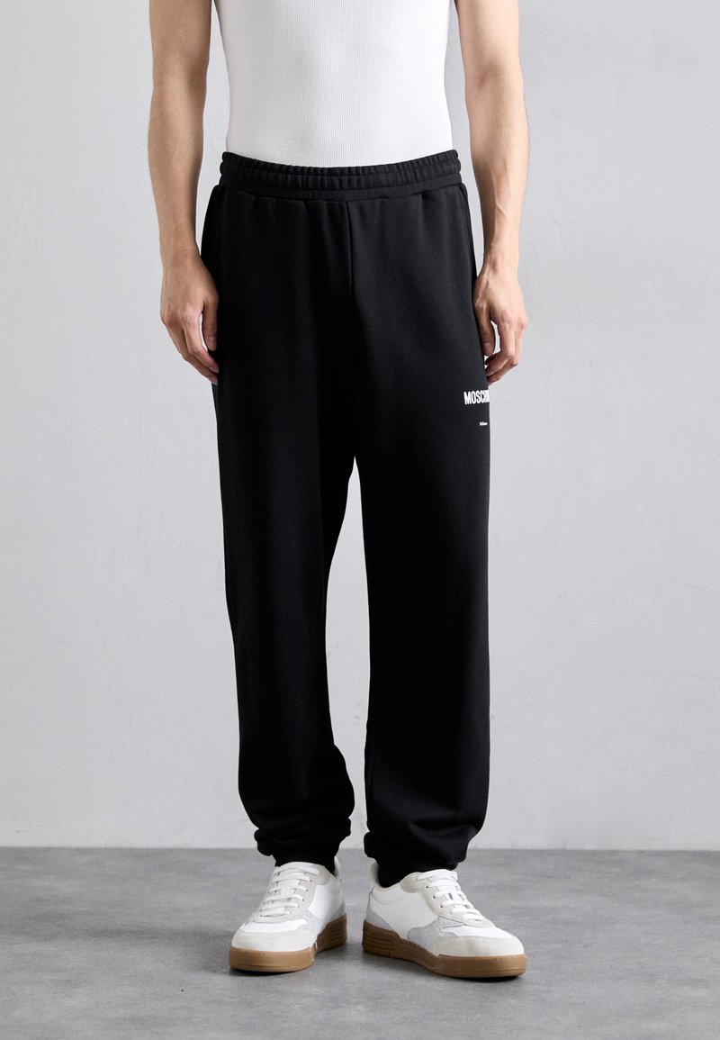 Svarta sweatpants med elastisk midja, sidofickor och vit logodetalj. Bärs med vita och grå sneakers. Minimalistisk design.