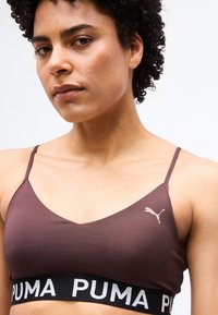 Γυναίκα με κοντά σγουρά μαλλιά που φοράει ένα καφέ αθλητικό σουτιέν Puma με λεπτές ράντες και μαύρη ταινία με λογότυπο.