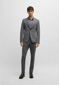 Grauer Anzug mit maßgeschneiderter Passform, aus glattem Stoff gefertigt. Verfügt über ein Zwei-Knopf-Jacket, Reverskragen und passende Slim-Fit-Hosen. Schwarze Schuhe.