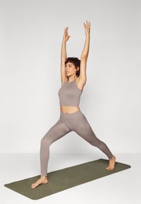 Light brown ribbed sports set with cropped top and high-waisted leggings. Model performs a yoga pose on a green mat against a neutral background.