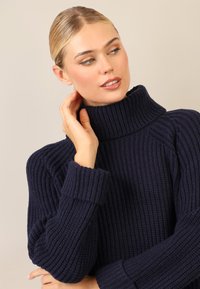 Maglione a coste di colore blu navy con colletto alto, polsini rifiniti e tessuto caratterizzato. Prima di un primo piano che mette in risalto il materiale e i dettagli di design.