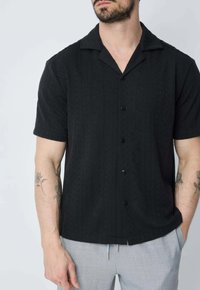 Chemise noire à manches courtes avec un motif texturé, col pointu et cinq boutons sur le devant, assortie à un pantalon gris clair.