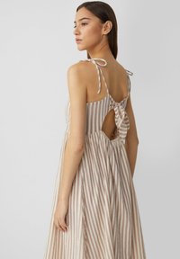 Gestreiftes Kleid mit dünnen Trägern, Schleifen-Detail am Rücken und einem ausgeschnittenen Element. Hergestellt aus leichtem Stoff, in beigen und sanften Rosatönen.