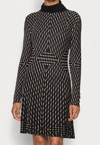 Frau trägt ein langärmliges Kleid mit schwarz-weißer Musterung, das einen hohen Ausschnitt und geometrische vertikale sowie horizontale Designs aufweist.