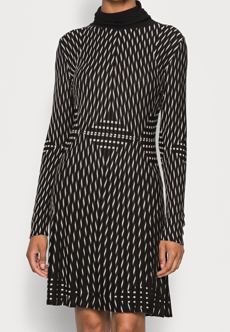 Frau trägt ein langärmliges Kleid mit schwarz-weißer Musterung, das einen hohen Ausschnitt und geometrische vertikale sowie horizontale Designs aufweist.