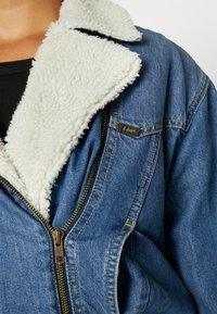 Kurtka jeansowa z białym kołnierzem sherpa, częściowo zamknięty mosiężny zamek, brązowa metka marki Lee na piersi, noszona na czarnym prążkowanym topie.