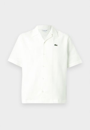 Īsām piedurknēm balta krekla kreklā ar faktūru, ar zaļu izšūtu logotipu kreisajā krūtīs un klasisku kempinga apkakli.