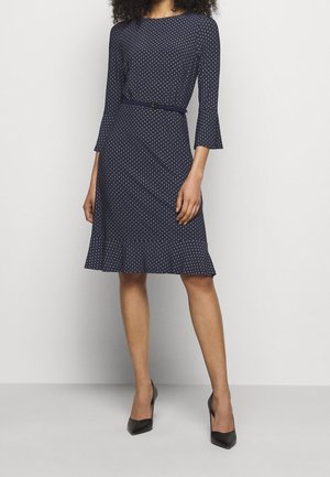 Nainen, joka pukeutuu tummansiniseen, polvenpituiseen mekkoon, jossa on pieniä valkoisia kuvioita, puolipitkät hihat, ohut vyö ja mustat teräväkärkiset korkokengät.