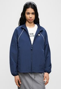 Ελαφρύ τζάκετ σε ναυτικό μπλε με φερμουάρ και υφή, με όρθιο γιακά και ελαστικές μανσέτες. Φορεμένο πάνω από λευκή μπλούζα.