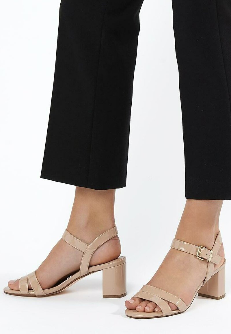 Sandales à talon bloc en cuir verni beige avec des lanières croisées, une sangle à la cheville avec boucle et une finition lisse et brillante.