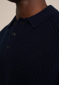 Marinblå stickad polo-tröja med en strukturerad mönster, med en ribbad krage och tre svarta knappar. Närbild.