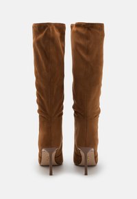 Botas altas de ante marrón con tacones delgados y altos, vistas desde la parte de atrás sobre un fondo blanco liso.