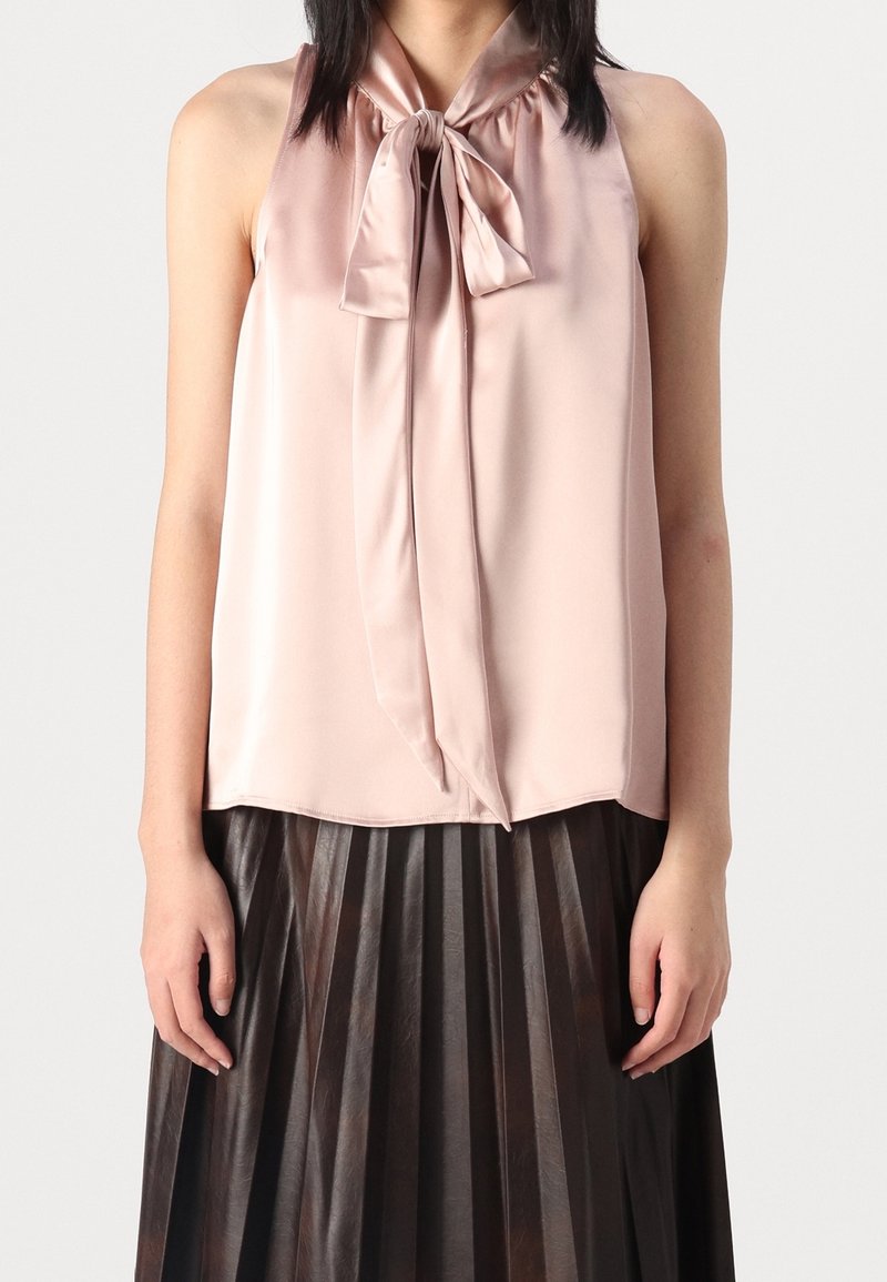 Donna che indossa una blusa senza maniche di satin rosa chiaro con un fiocco al collo e una gonna plissettata alta di colore marrone scuro, su uno sfondo neutro.