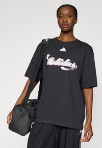 Μαύρο φαρδύ μπλουζάκι με λευκό και ροζ λογότυπο "Adidas". Το μοντέλο κρατά μια μικρή μαύρη αθλητική τσάντα με ιμάντα ώμου. Απλό φόντο.