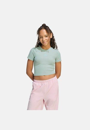 Giovane donna con capelli ricci che indossa una maglietta corta verde chiaro e pantaloni rosa larghi, in piedi con le mani dietro la schiena.