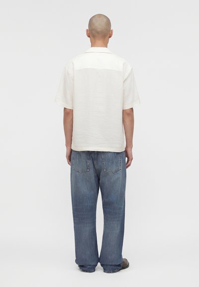 Άνδρας με ξυρισμένο κεφάλι κοιτώντας προς τα πίσω, φοράει λευκό κοντομάνικο πουκάμισο και φαρδιά μπλε τζίν against a plain light background.